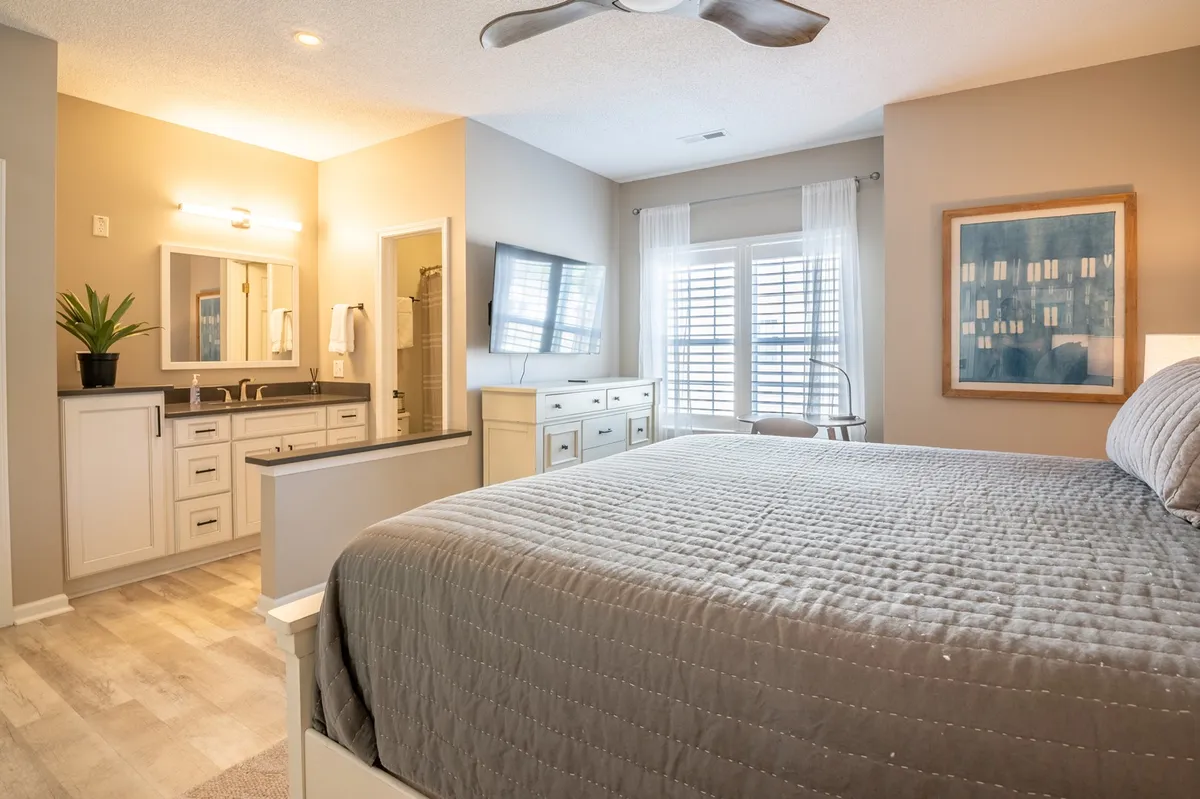 A tranquil king suite featuring a plush king-sized bed with fresh linens, a bathroom vanity in view, a large window with shutters allowing natural light, and tasteful wall art. The suite includes a Smart TV for entertainment and a ceiling fan for comfort, encapsulating the core amenities described.
