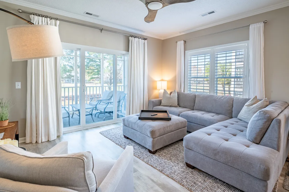 A welcoming living area with a large sectional sofa, coffee table, floor lamp, and a sliding glass door that opens to an outdoor view. The abundance of natural light and direct connection to the exterior space echo the feel of a porch that bridges inside comfort with nature. The view beyond hints at greenery, reinforcing the sense of place within a golf community.