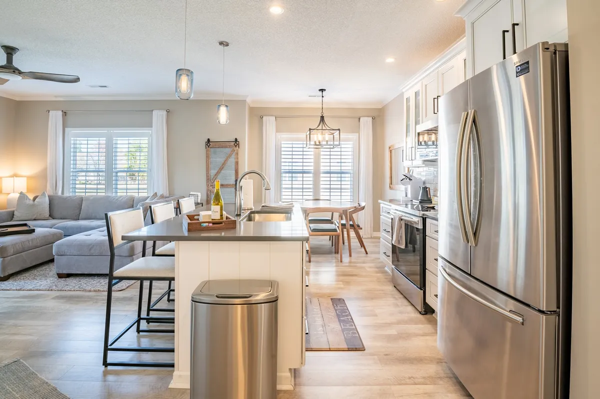 A modern kitchen with a spacious island countertop and seating, stainless steel appliances, large windows, and an adjoining dining area. The layout encourages entertaining and seamless movement between cooking, dining, and conversation.