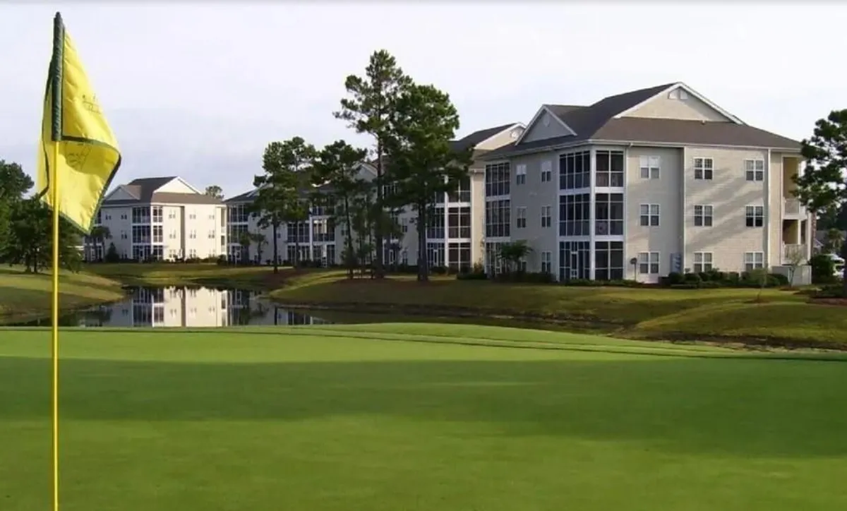 An exterior view showing the golf flag, tranquil water with reflections, surrounding trees, and residential buildings in the background. The peaceful mood and sunset-ready orientation evoke the scenic fairway vistas described in the section, emphasizing both the location and the atmosphere the porch overlooks.