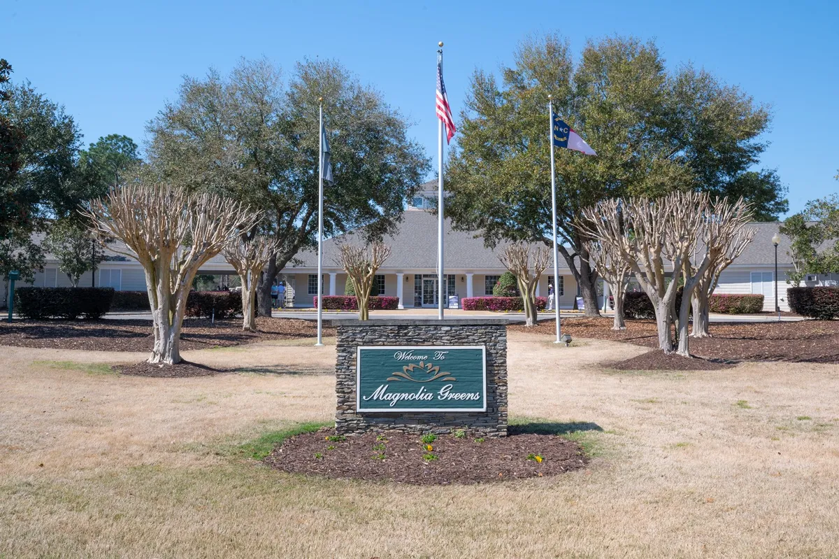 This exterior image features a welcoming entrance with a prominent 'Welcome' sign, flagpoles, neatly trimmed trees, and the inviting facade of a community building. The landscaping and open space suggest an environment suitable for gatherings and outdoor events, hinting at the lively community and accessible amenities nearby.