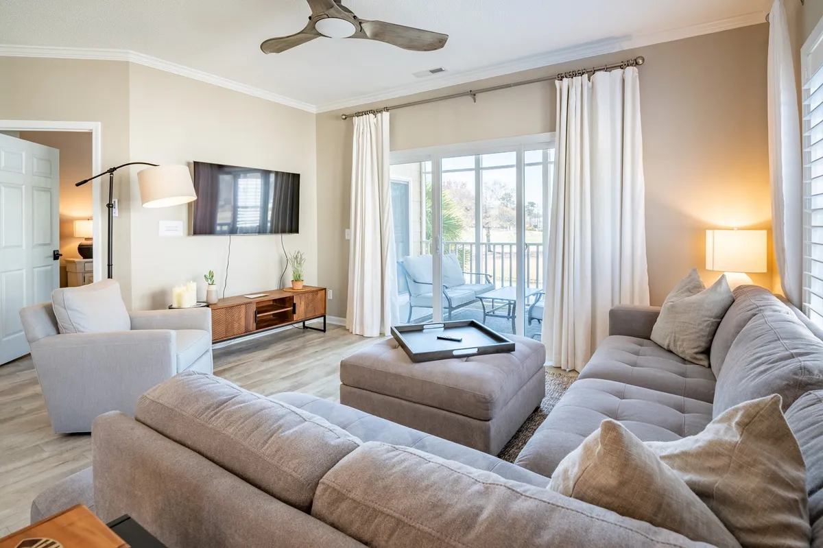 This image presents a cozy, sunlit living room featuring a comfortable sectional sofa, a plush armchair, a modern coffee table, and large windows that bathe the space in natural light. The atmosphere is both inviting and ideal for relaxation or social gatherings.
