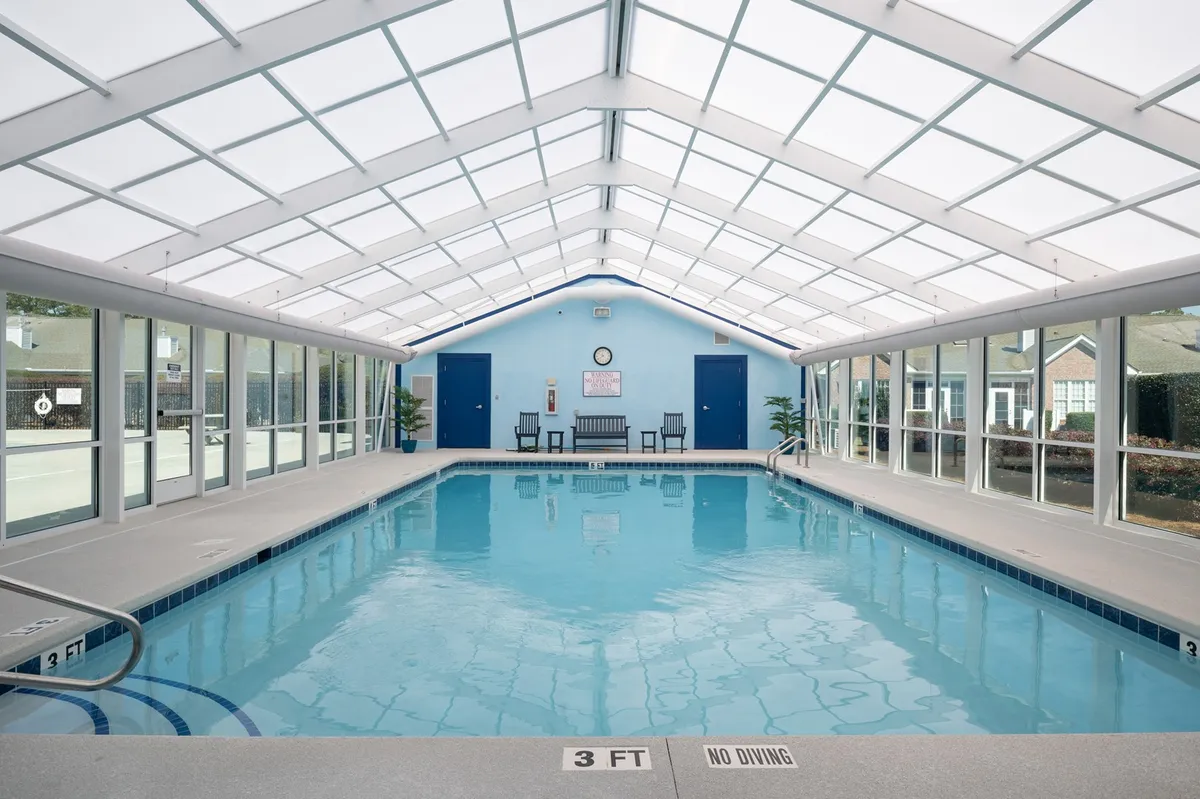A bright indoor pool area with a glass ceiling that allows sunlight to pour in, lined with comfortable lounge chairs and vibrant blue walls. The setting is calm and inviting, perfect for morning swims or gathering with friends on rainy days.