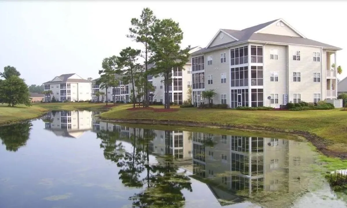 A peaceful waterfront scene featuring apartment buildings tucked among mature trees, with calm water reflecting the sky and foliage. The area invites leisurely strolls, sunset watching, and moments of tranquility amid nature.
