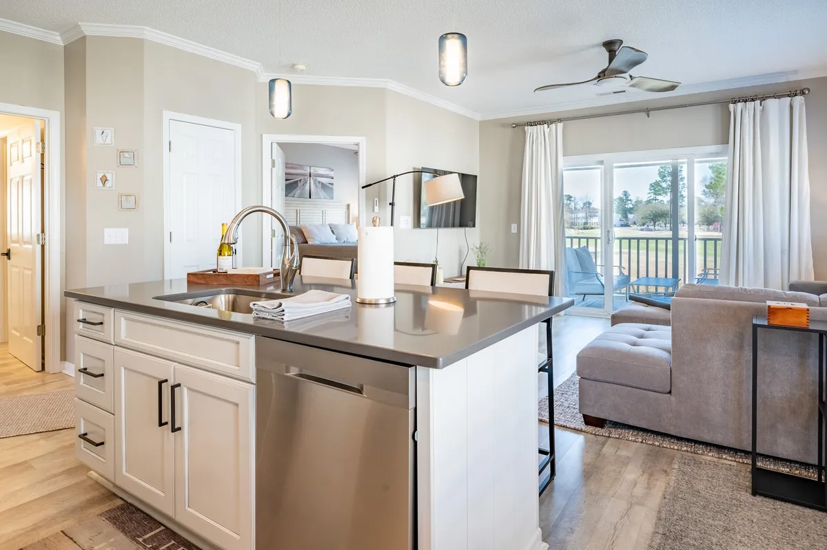 A bright, open-plan living space featuring a kitchen island that flows into a comfortable living area. Large windows invite in natural light, and offer views to the balcony, hinting at the outdoor environment beyond. The scene suggests opportunities for relaxing, entertaining, and enjoying the surrounding scenery from inside the condo.