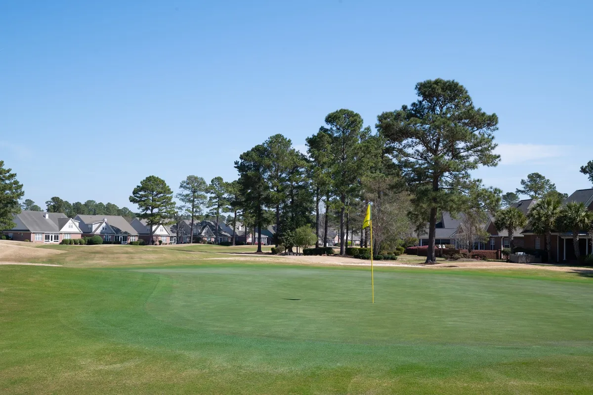 A peaceful scene of the golf green, complete with a yellow flag, surrounded by mature trees and residential buildings. The image captures the quiet elegance of the course and hints at the convenience of resort living adjacent to world-class golfing.