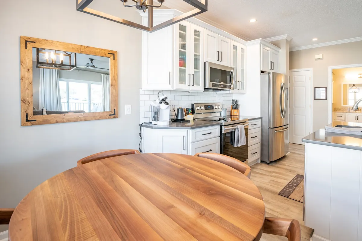 A warm and inviting dining area with a wooden table set for four, modern kitchen appliances in the background, and a framed mirror that enhances the sense of space. This spot is designed for shared meals and vibrant conversation.