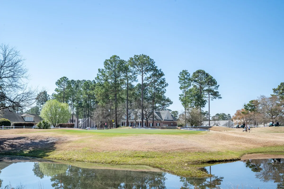 A wide view of Magnolia Greens’ championship golf course, featuring lush fairways bordered by a serene pond, pine trees, and glimpses of residential buildings in the background. The well-kept greens and natural landscaping highlight the inviting atmosphere for golfers and residents alike.